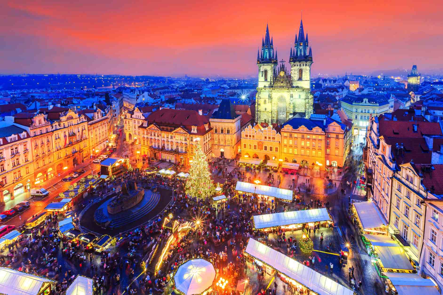 Prague Old Town Square Christmas market at twilight, featuring festive stalls, illuminated historic buildings, and the Gothic towers of the Church of Our Lady before Týn.